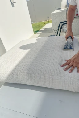 Proceso de limpieza profunda de un cojín blanco con aspiradora para eliminar manchas y suciedad. Limpieza profesional de cojín blanco con aspiradora en un espacio exterior iluminado.