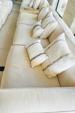 Sofá elegante con cojines de diseño que complementan una sala moderna y luminosa. Sofá beige con cojines decorativos blancos y rayas marrones en una sala luminosa.